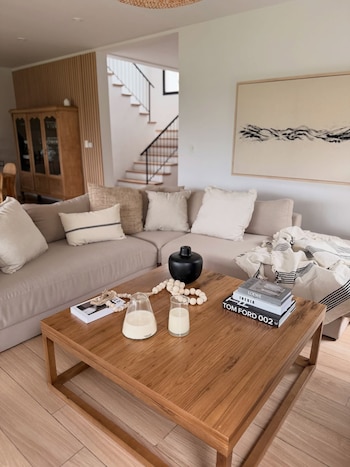 Salón moderno con sofá beige en L, cojines y manta a rayas. Mesa de centro de madera clara con jarrón negro, cuentas decorativas, velas y libros. Escalera en el fondo