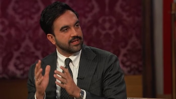 Hombre con barba, traje oscuro y corbata que gesticula con las manos frente a un fondo de color granate