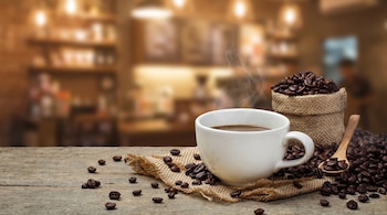 Una taza de café caliente con vapor, un saco de arpillera con granos de café y granos sueltos sobre una mesa de madera. Fondo borroso de cafetería.