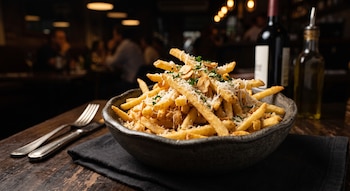Plato hondo de papas fritas con queso rallado, láminas de ajo y perejil picado en una mesa de madera. Detrás, botellas de vino y aceite, y personas borrosas