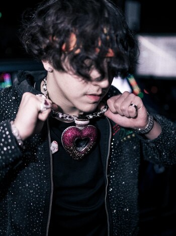 Persona con cabello oscuro, chaqueta con brillos, collar de joyas con dije de corazón, anillos y pulsera, ajustando el collar