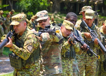 Varios soldados con uniformes de camuflaje y gorras están de pie al aire libre, sosteniendo fusiles negros. Algunos llevan chalecos tácticos verdes