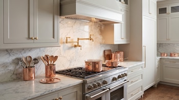 Una cocina con gabinetes superiores e inferiores grises, encimeras de mármol blanco veteado, una estufa de acero inoxidable y utensilios de cobre.