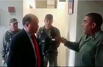 Primer plano de hombre en traje y corbata roja frente a hombre con camisa verde que extiende su mano, flanqueados por dos guardias en uniforme oscuro