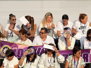 Personas vestidas de blanco sentadas en gradas sostienen estampas religiosas y papeles para protegerse del sol ante una pancarta morada
