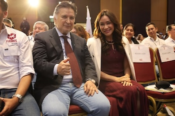 Un hombre y una mujer sentados, el hombre de traje y el pulgar arriba, la mujer con vestido y blazer. Sillas rojas con letreros "RESERVADO". Un logo FIRMES en camisa
