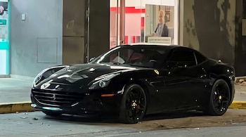 Un automóvil Ferrari negro estacionado en la calle frente a un edificio con paneles de vidrio y un cartel publicitario interno.