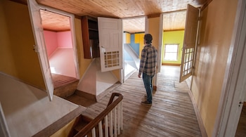 Un hombre de espaldas en un pasillo con suelo y techo de madera, mirando habitaciones de paredes rosa, amarillo y azul; una escalera de madera a la izquierda.
