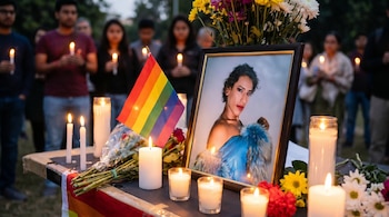 Primer plano de un altar con velas, flores, una bandera LGBTQ+ y el retrato de una persona con cabello rizado. Personas sosteniendo velas de fondo.