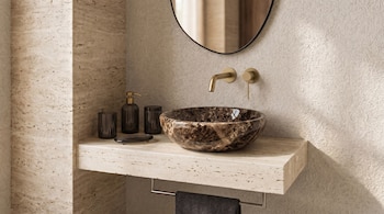 Lavabo de mármol, grifo dorado en la pared, encimera de travertino, dispensador de jabón, vasos, jabonera, toalla oscura, espejo ovalado, pared texturizada.