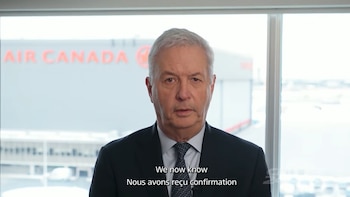 Un hombre de mediana edad con cabello gris y traje oscuro habla frente a una ventana, con el logo 'AIR CANADA' visible en un edificio al fondo