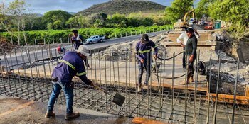 Varios obreros con palas y herramientas trabajan sobre una red de varillas de acero junto a una carretera y una excavadora