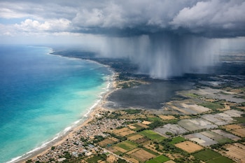 Vista aérea de la costa peruana con una gran cortina de lluvia sobre el mar y la tierra, zonas urbanas junto a la playa y campos agrícolas inundados.