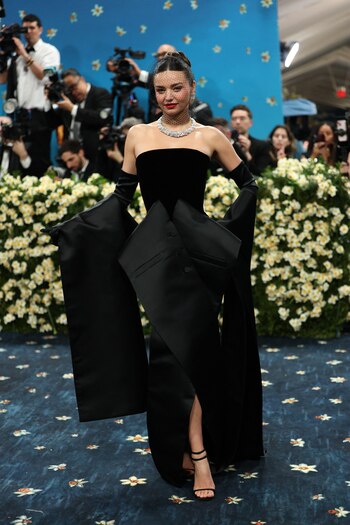 Miranda Kerr usó un vestido de Dior: negro de terciopelo sin tirantes, de silueta estructurada y volumen escultórico en la parte inferior, generado por pliegues asimétricos y una aplicación a modo de lazo XL con botones forrados. El diseño mostró una abertura frontal que dejó al descubierto una pierna, y se complementó con guantes largos de satén que terminaban en mangas acampanadas y alargadas que colgaban hasta el suelo