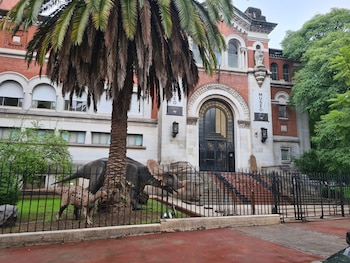 Vista frontal de un edificio museo de ladrillo rojo con una palmera Babassu alta, y estatuas de dinosaurios, incluyendo un triceratops, detrás de una valla