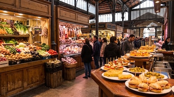 Interior de un mercado con puestos de verduras, embutidos y platos de tapas, aceitunas y tortilla de patatas con personas en el fondo.