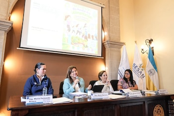 Personas reunidas en un salón con una mesa directiva, un orador en un podio, un intérprete de lengua de señas, banderas y una pantalla