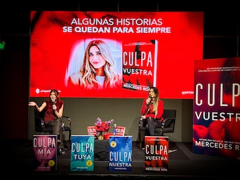 Dos mujeres sentadas en un escenario con micrófonos, frente a una pantalla roja con una imagen de la autora y la portada de "Culpa Vuestra". Varios libros de la saga "Culpa" están en el suelo