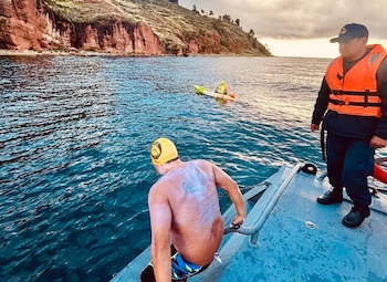 Un nadador con gorro amarillo y crema en la espalda se prepara para entrar al agua desde un barco. Un oficial y un kayak lo asisten, con una orilla rocosa de fondo
