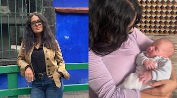 Mujer con gafas, chaqueta color camel y jeans, apoyada en una valla verde. Otra persona de cabello oscuro sostiene a un bebé dormido vestido de blanco.