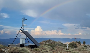 Una estación de monitoreo con panel solar y antenas en la cima de una montaña, con un arcoíris visible en el cielo