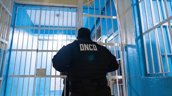 Un hombre de espaldas con uniforme negro y las letras DNCD en la espalda, dentro de un espacio con rejas blancas y paredes azules.