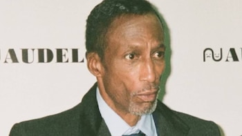 Un hombre afroamericano con traje oscuro y barba canosa posa ante un panel claro con la palabra AUDELCO