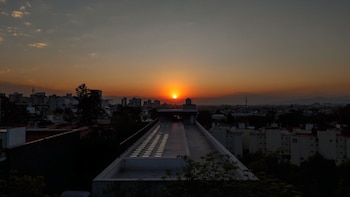 Vista panorámica de un amanecer con el sol naranja brillante sobre un horizonte urbano con edificios y montañas a lo lejos, el primer plano muestra el techo de un edificio moderno