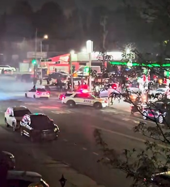 Vista aérea de una calle concurrida de noche, donde un coche de policía con luces intermitentes está entre varios coches y personas, con humo en el aire