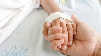 Primer plano de una mano adulta sosteniendo la mano de un niño. La mano del niño tiene una vía intravenosa con esparadrapo sobre una sábana con diseño azul.