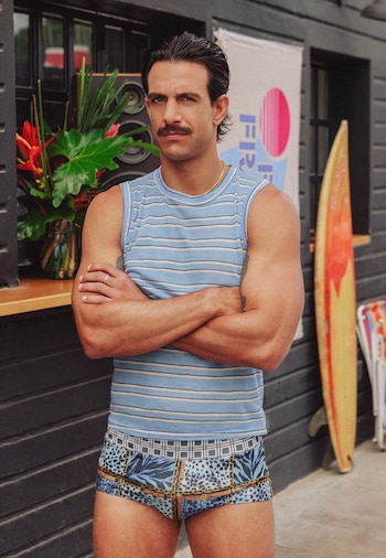 Hombre con bigote y cabello oscuro, camiseta de rayas azules y blancas, bañador con estampado animal y cadenas, brazos cruzados. Tabla de surf al fondo