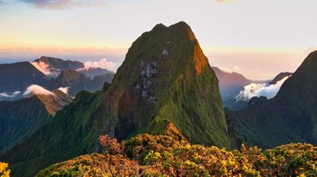 Vista panorámica de una gran montaña verde con otras montañas y nubes, cielo claro y vegetación en primer plano.