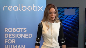 Robot humanoide con cabello rubio, traje blanco y brazos robóticos negros, de pie frente a un banner azul con el logo de Realbotix y pantallas de fondo