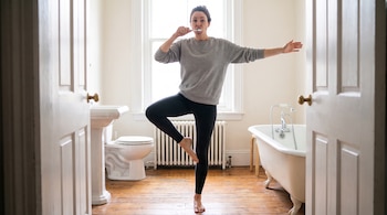 Mujer de pie sobre una pierna con los brazos extendidos, cepillándose los dientes en un baño con inodoro, lavamanos y bañera, enmarcada por dos puertas abiertas.