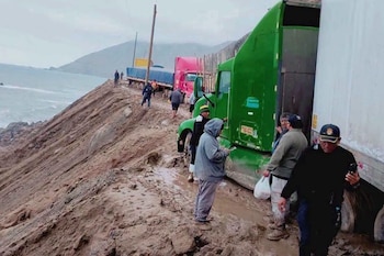 Camiones de carga y personas detenidos sobre un tramo cubierto de lodo en un camino costero junto a un acantilado