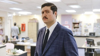 Primer plano de Fernando Bonilla con bigote, traje azul a cuadros, camisa blanca y corbata negra, mirando fijamente en un entorno de oficina