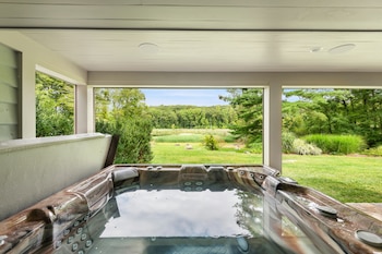 Jacuzzi en una terraza cubierta con agua clara, reflejando el cielo. A través de una ventana, se ve un paisaje verde con árboles, un estanque y cielo azul