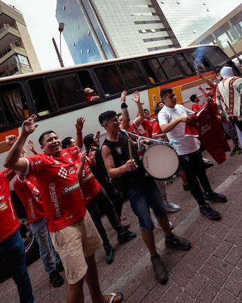 Un grupo de personas vestidas con camisetas rojas alza los brazos y toca un tambor junto a un autobús en la vía pública