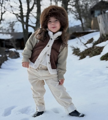 Un niño con gorro de piel marrón, chaqueta beige, chaleco marrón con ribete de piel blanco y pantalones claros de pana, de pie en un paisaje nevado con árboles