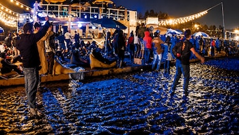 Imagen nocturna de un festival al aire libre con un suelo cubierto de barro y charcos, donde la gente camina entre luces de guirnalda y estructuras de madera