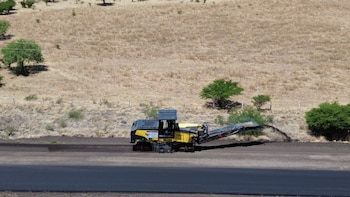 Una máquina de pavimentación amarilla y negra sobre una carretera oscura, con material procesado saliendo, flanqueada por una ladera árida con arbustos verdes