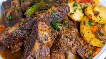 Un plato con trozos grandes de carne de res estofada, papas cortadas en rodajas, cebolla, zanahoria, hojas de laurel y perejil fresco picado.