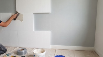 Mano de persona aplicando adhesivo con espátula sobre paneles blancos de poliestireno en una pared. Cubeta con pasta, rollo de malla y herramientas en el suelo.