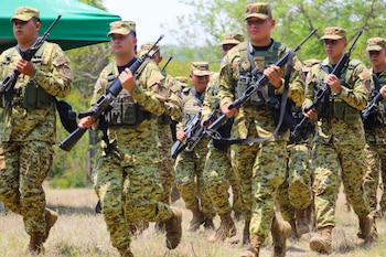 Un grupo de soldados con uniformes de camuflaje, chalecos tácticos y rifles avanza sobre hierba seca. Un toldo verde se ve a la izquierda y árboles de fondo
