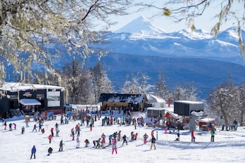 Grupo de personas Mapuche alrededor de una fogata, con un tambor, frente a pistas de esquí nevadas y un remonte en el Cerro Chapelco, bajo un cielo azul. Una bandera Mapuche flamea a la derecha