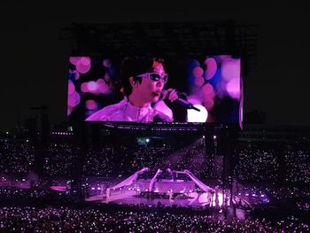 Pantalla gigante con un cantante con gafas oscuras sobre un escenario en un estadio nocturno lleno de puntos de luz morados