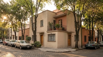 Vista exterior de una casa de tonos neutros en una calle arbolada de la Ciudad de México, con coches aparcados y luz cálida de atardecer filtrándose entre los árboles.