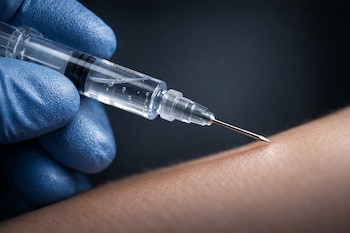 Una mano con guante azul sujeta una jeringa transparente con líquido incoloro. La aguja de la jeringa apunta al brazo de una persona.