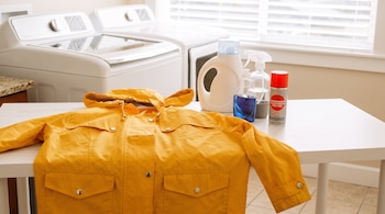 Una chaqueta amarilla extendida sobre una mesa blanca junto a botellas de detergente, rociadores y un aerosol impermeabilizante.