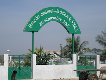 Un arco verde con letras blancas ante la escultura de un ancla, con una verja blanca y vegetación de fondo